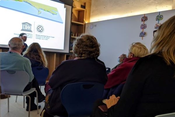 A man is giving a presentation to a room full of seated people. The presentation screen shows an image of a coastal map with a UNESCO symbol beneath it. 